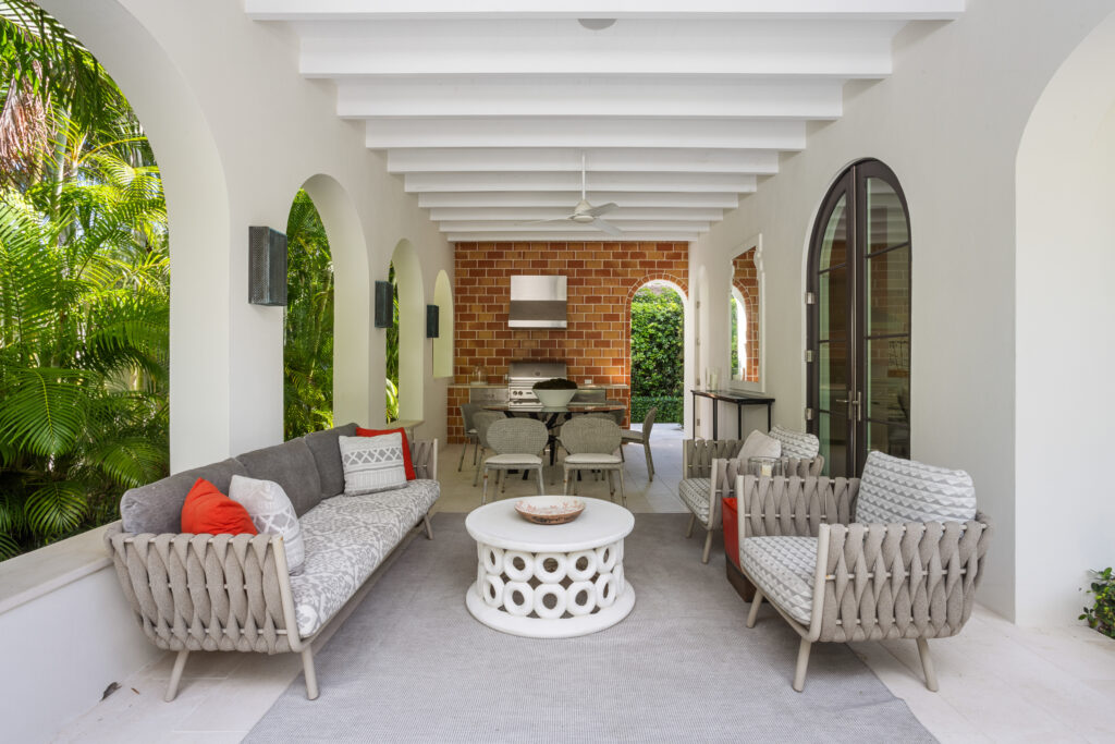 Patio with cushioned woven sofas and armchairs around a round white coffee table, featuring a dining area and an outdoor kitchen with a grill against a brick wall, under a white ceiling with exposed beams and arched openings revealing lush greenery.