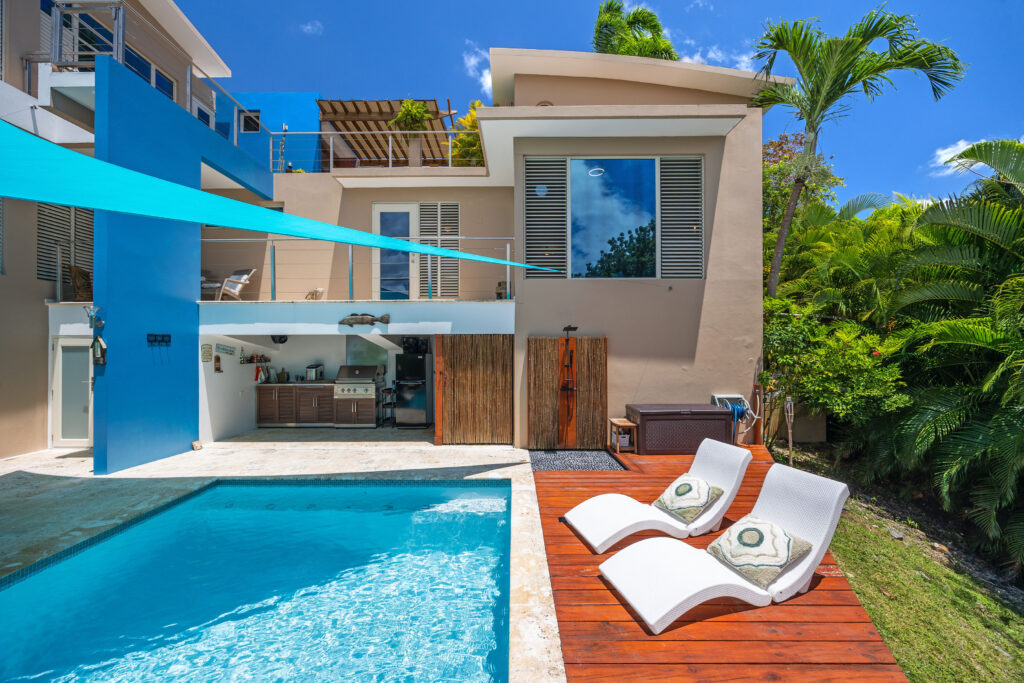 Modern outdoor area with a swimming pool, white lounge chairs on a wooden deck, and an outdoor kitchen and dining area on the ground floor of a multi-level house, shaded by blue sails and surrounded by lush greenery.