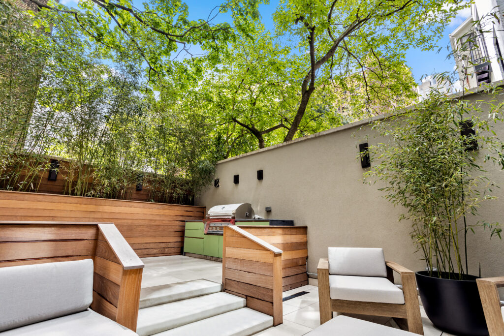 Outdoor patio area with wooden chairs and a bench, featuring a built-in grill and countertop against a wall, surrounded by trees and bamboo plants under a clear blue sky.