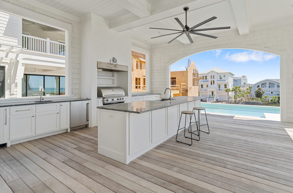 Modern outdoor kitchen with white cabinetry, a granite island with two bar stools, built-in stainless steel grill and sink, wooden flooring, and an open ceiling with exposed beams and a ceiling fan, overlooking neighboring houses and a pool.
