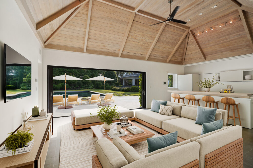 Modern pool house interior with beige sectional sofas, blue and beige pillows, a wooden coffee table, and sliding glass doors opening to a patio with lounge chairs, umbrellas, and a pool; a kitchen with a white island and wooden bar stools is also visible.
