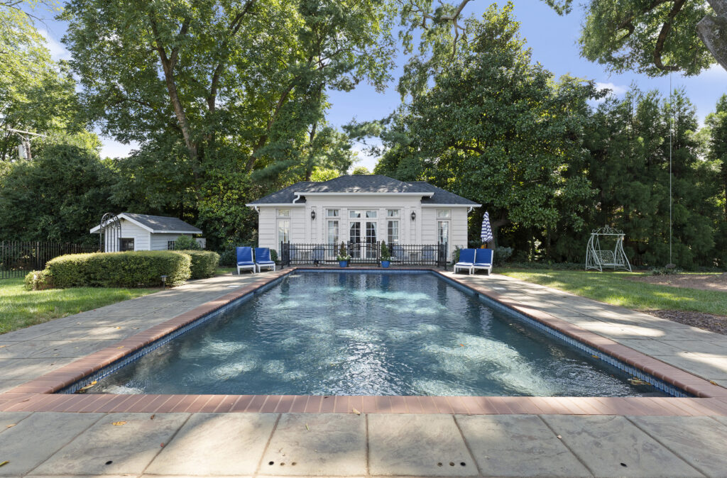 Rectangular swimming pool with clear blue water, a small white pool house with large windows at the far end, blue lounge chairs, an umbrella, surrounding greenery, and a swing set to the right.
