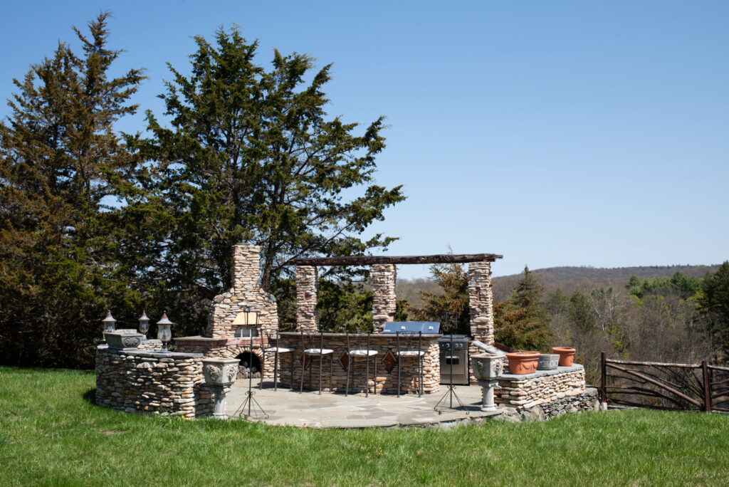 Bluestone patio with a pizza oven, grill, and bar, surrounded by grass and trees.