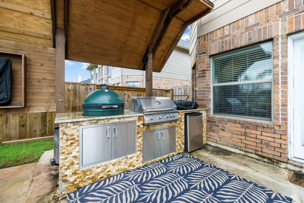 Outdoor kitchen area with a covered wooden roof, featuring a green egg grill, a stainless steel gas grill, and a mini fridge.