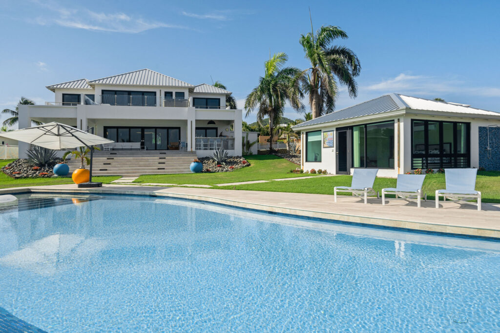 Luxurious property with a large swimming pool, a modern two-story house with a balcony, a smaller pool house, green lawns, palm trees, and poolside lounge chairs with an umbrella.