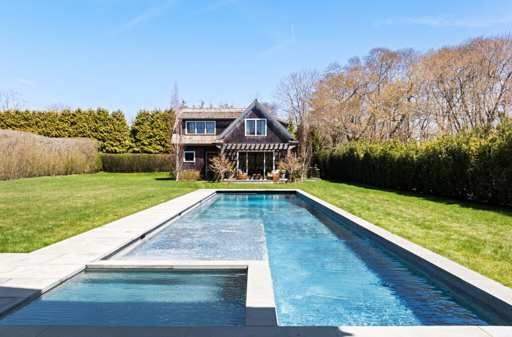 Cottage-like pool house with large windows and an outdoor seating area under a pergola, situated behind a rectangular swimming pool with a shallow section, surrounded by grass, hedges, and trees under a clear sky.