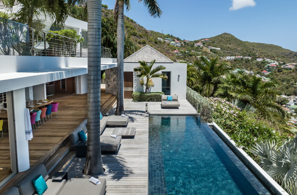 A modern house with an open-plan design overlooks an infinity pool surrounded by wooden decking, lounge chairs, and palm trees, with hills and clear blue skies in the background.