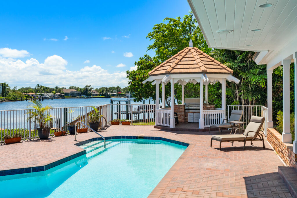 Outdoor scene with a swimming pool surrounded by a brick patio, two lounge chairs, a white gazebo with a brown shingled roof, and a view of a body of water with houses and trees on the opposite shore under a blue sky.