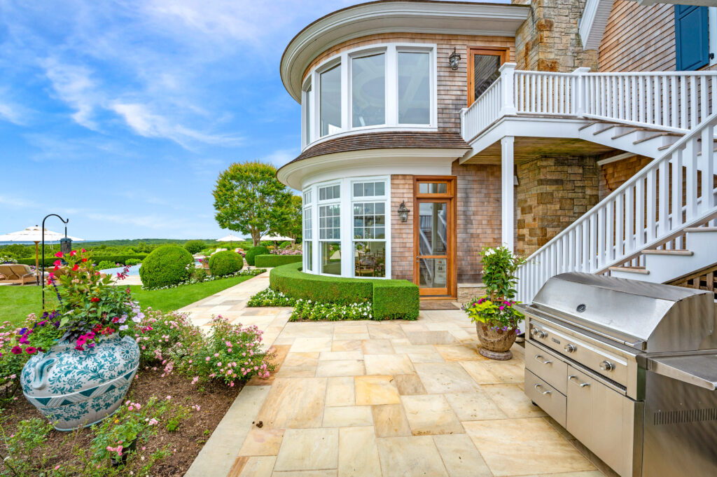 Outdoor kitchen patio with a stainless steel barbecue grill, surrounded by lush landscaping, colorful flowers, and an expansive lawn with seating and umbrellas.