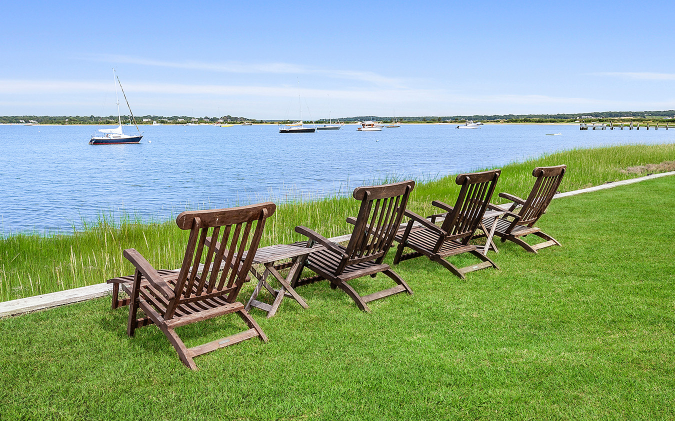 Four wooden lounge chairs on a grassy lawn face a calm harbor with anchored boats under a clear blue sky.