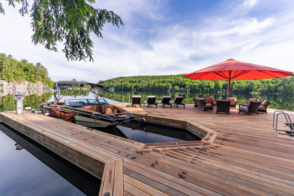 A wooden dock with lounge chairs and a red umbrella extends over calm water, with a motorboat moored nearby and lush green trees and rocky cliffs in the background under a partly cloudy sky.