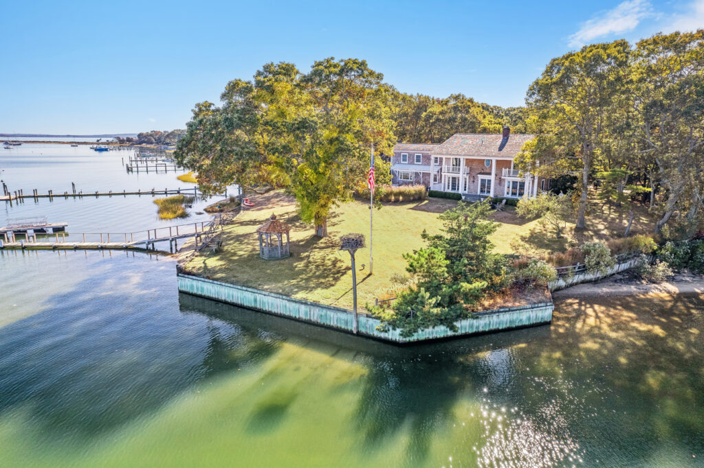 A large wooden house sits on a grassy, tree-lined property that extends into calm water, bordered by a wooden retaining wall, with docks and boats visible in the background under a clear blue sky.