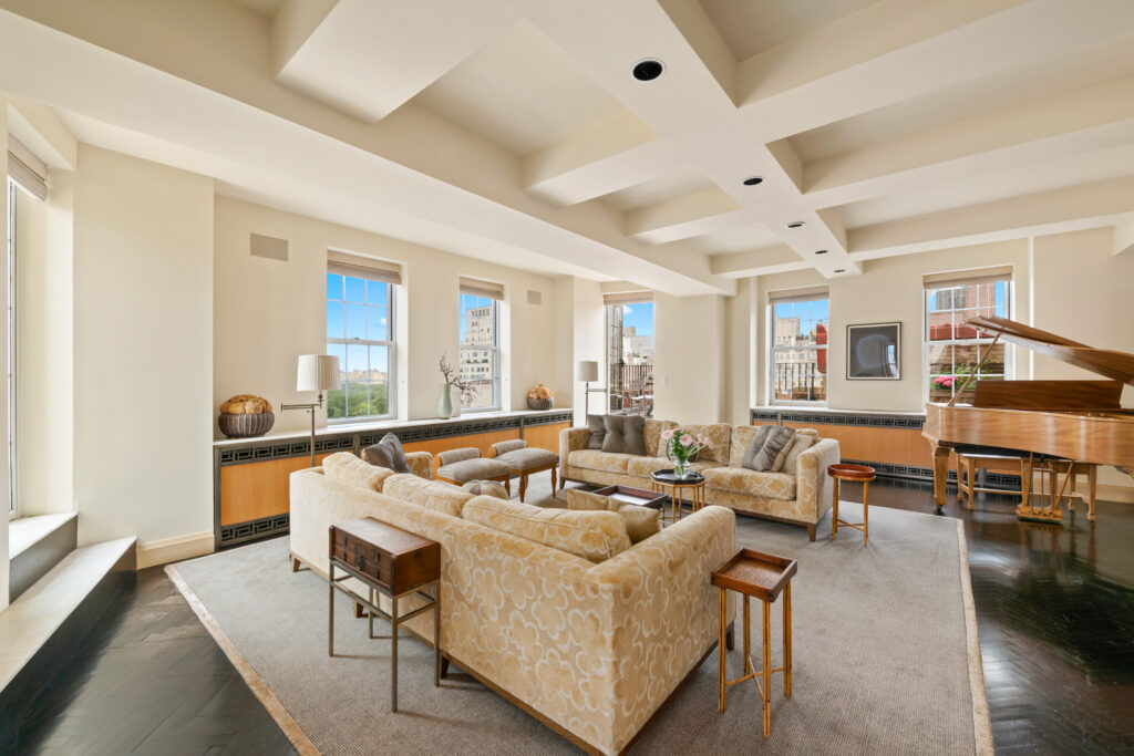 28 East 70_17 A spacious, well-lit penthouse living room with beige U-shaped sofas around a central coffee table, large windows showing a cityscape, a grand piano in the corner, decorative vases and plants on the window sills, and a ceiling with recessed lighting.