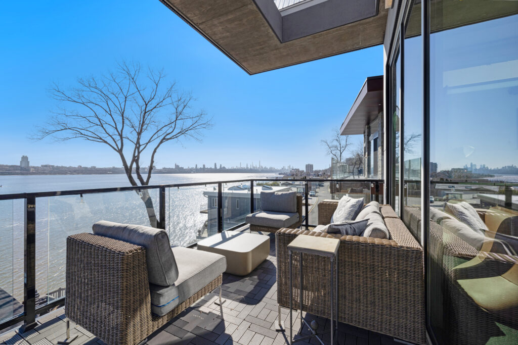 A modern patio with wicker furniture and glass railings overlooks a body of water, with bare trees and distant city buildings visible under a clear blue sky.