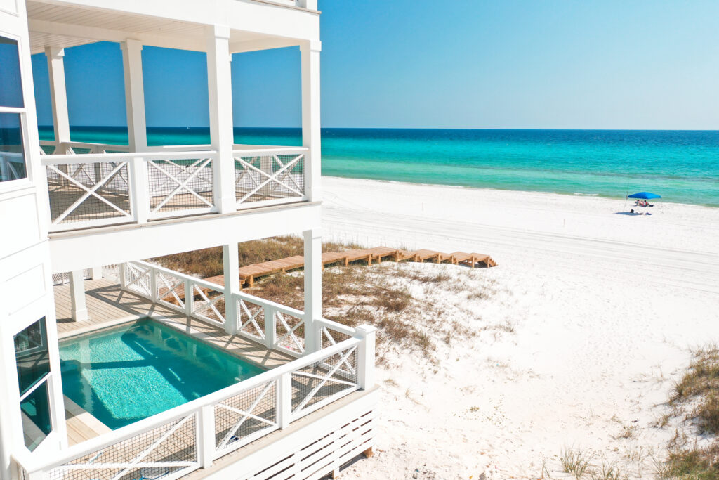 A beachfront house with white railings and multiple balconies features a private pool on the lower level, overlooking a white sandy beach and turquoise ocean under a clear blue sky.