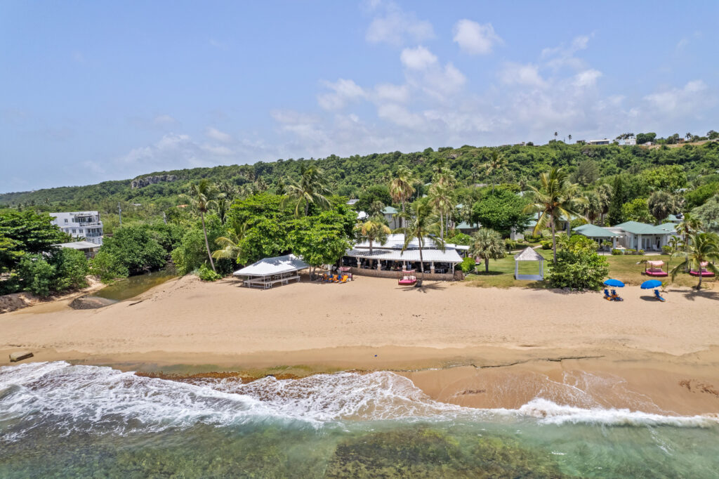 A tropical beachfront scene with gentle ocean waves meeting a sandy shore lined with colorful umbrellas and lounge chairs, backed by white tents and buildings nestled among lush green trees.