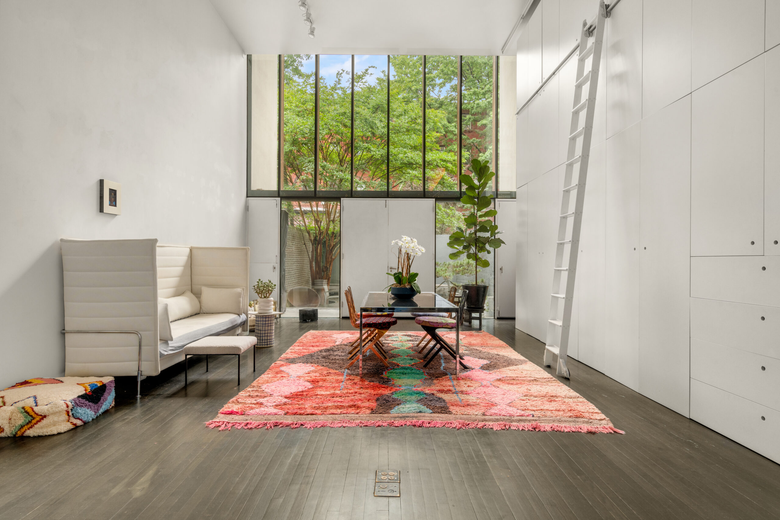 A modern living room with a high ceiling, large windows showcasing greenery outside, a white sofa, colorful rug, glass coffee table with plants, and a tall ladder leading to upper storage cabinets.