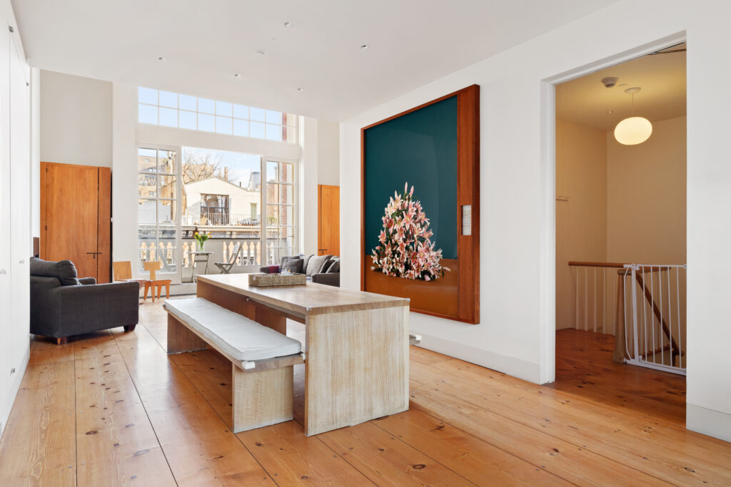 52 West 9th Street, Greenwich Village A modern, spacious penthouse dining area with a long wooden table and benches, large windows letting in natural light, a gray sofa in the background, and a large floral artwork on the wall.