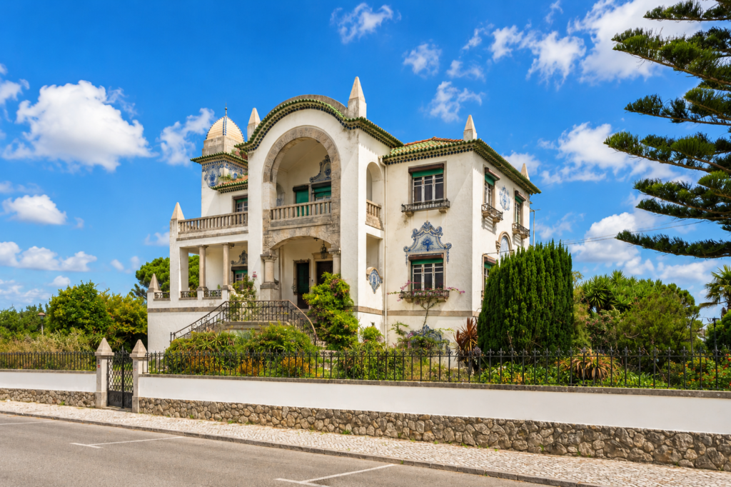 A large, ornate white villa with green trim, arched entry, tile murals, and lush gardens basking in the radiant energy of leo season under a bright blue sky with scattered clouds. A stone fence and gate separate the villa from the street.