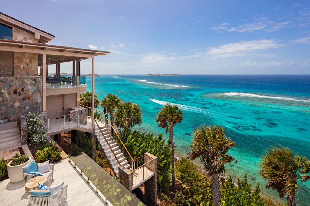A luxurious waterside home with a stone exterior and tiered terraces featuring outdoor seating and lush greenery, overlooking a turquoise ocean with coral reefs and distant small islands.