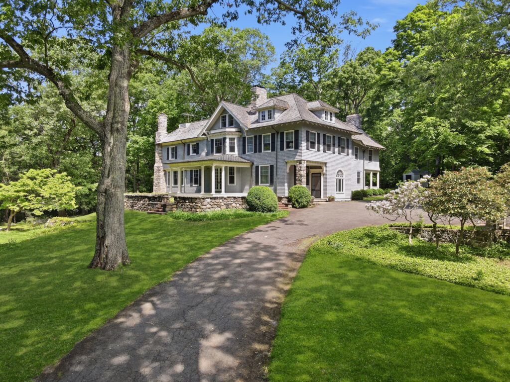 Large gray two-story house with white trim, stone chimney, and multiple windows, surrounded by a green lawn, trees, and shrubs, with a curved paved driveway leading to the entrance. The bright, sunny day captures the vibrant energy of leo season.