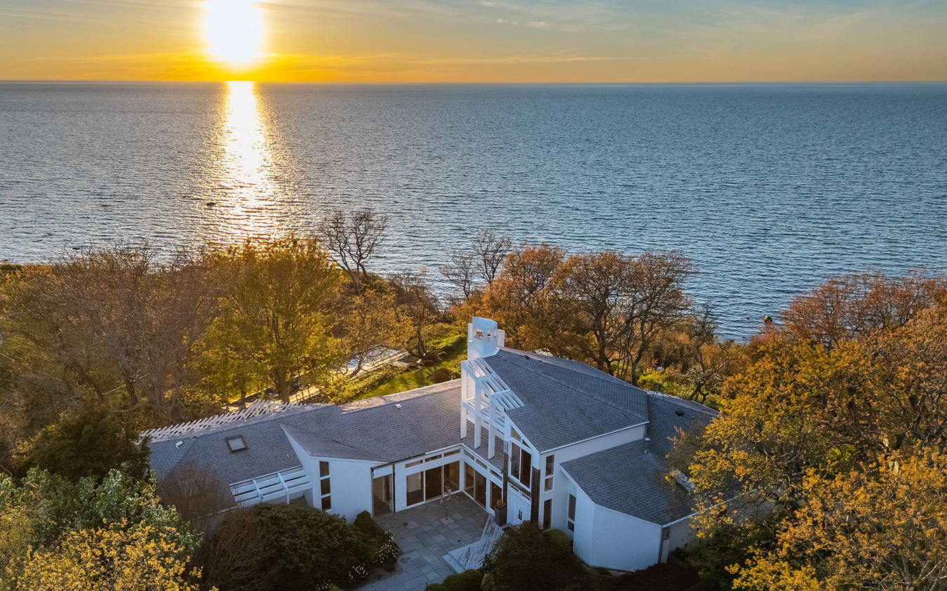 A modern waterside home with a gray roof sits on a tree-lined cliff overlooking a large body of water, with the sunset casting a golden reflection across the surface.