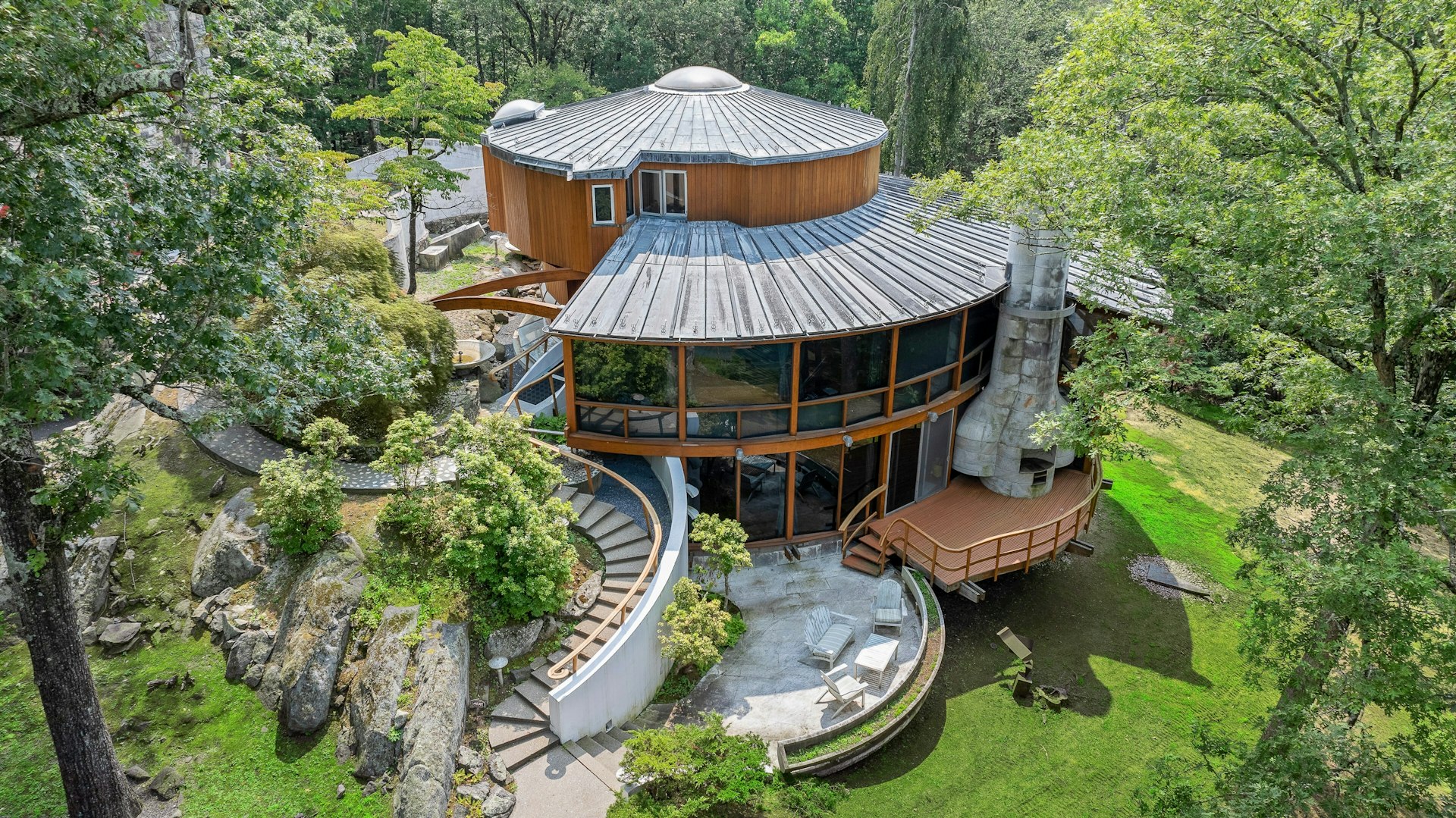 Aerial view of a unique, round modern house with large glass windows, a curved staircase, and surrounded by lush green trees—an architectural gem set to be featured on the A-list: September 2025. Wood, stone, and metal elements highlight its design.