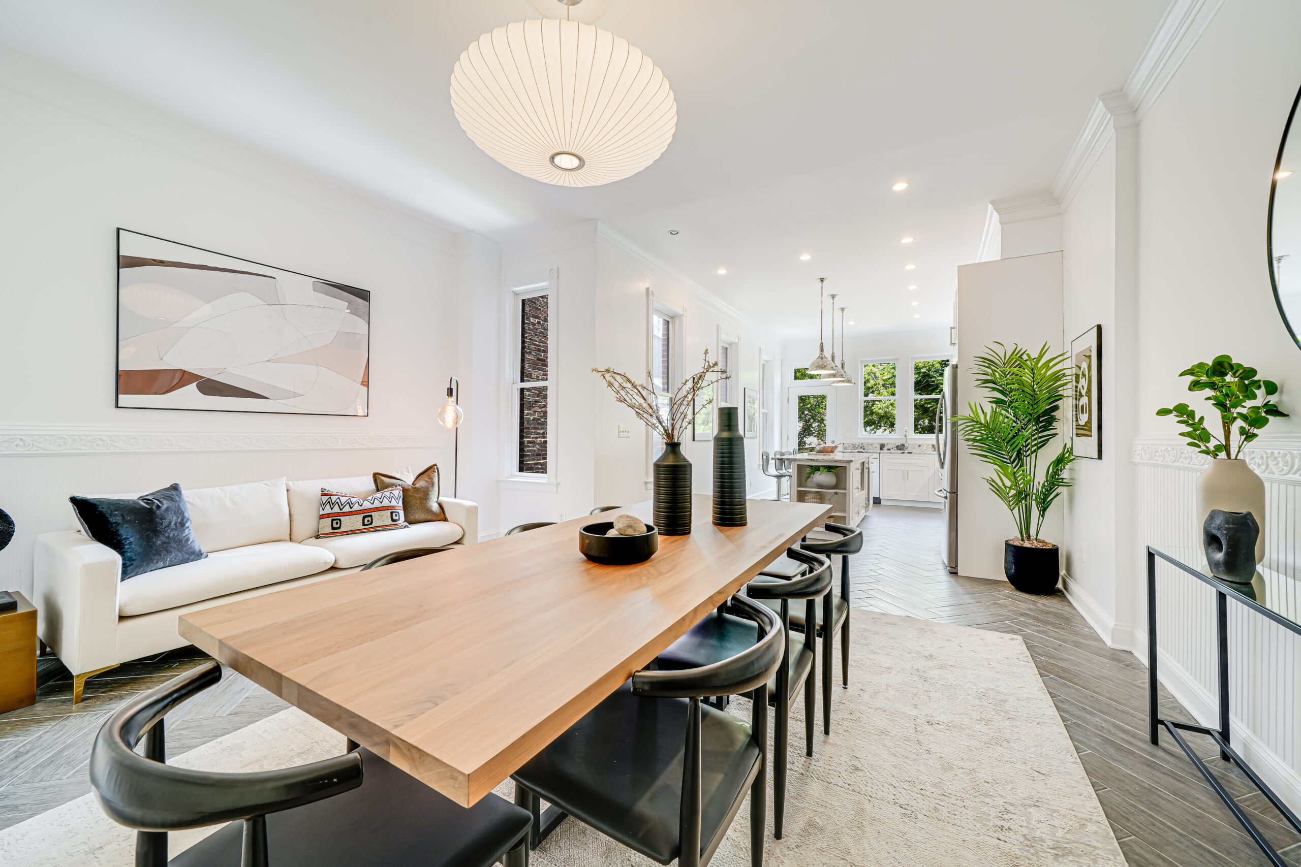 Modern open-concept living and dining area with a wooden table, black chairs, white sofa, abstract wall art, large pendant light, and greenery; kitchen visible in the background—perfect for hosting your A-list: September 2025 gatherings.