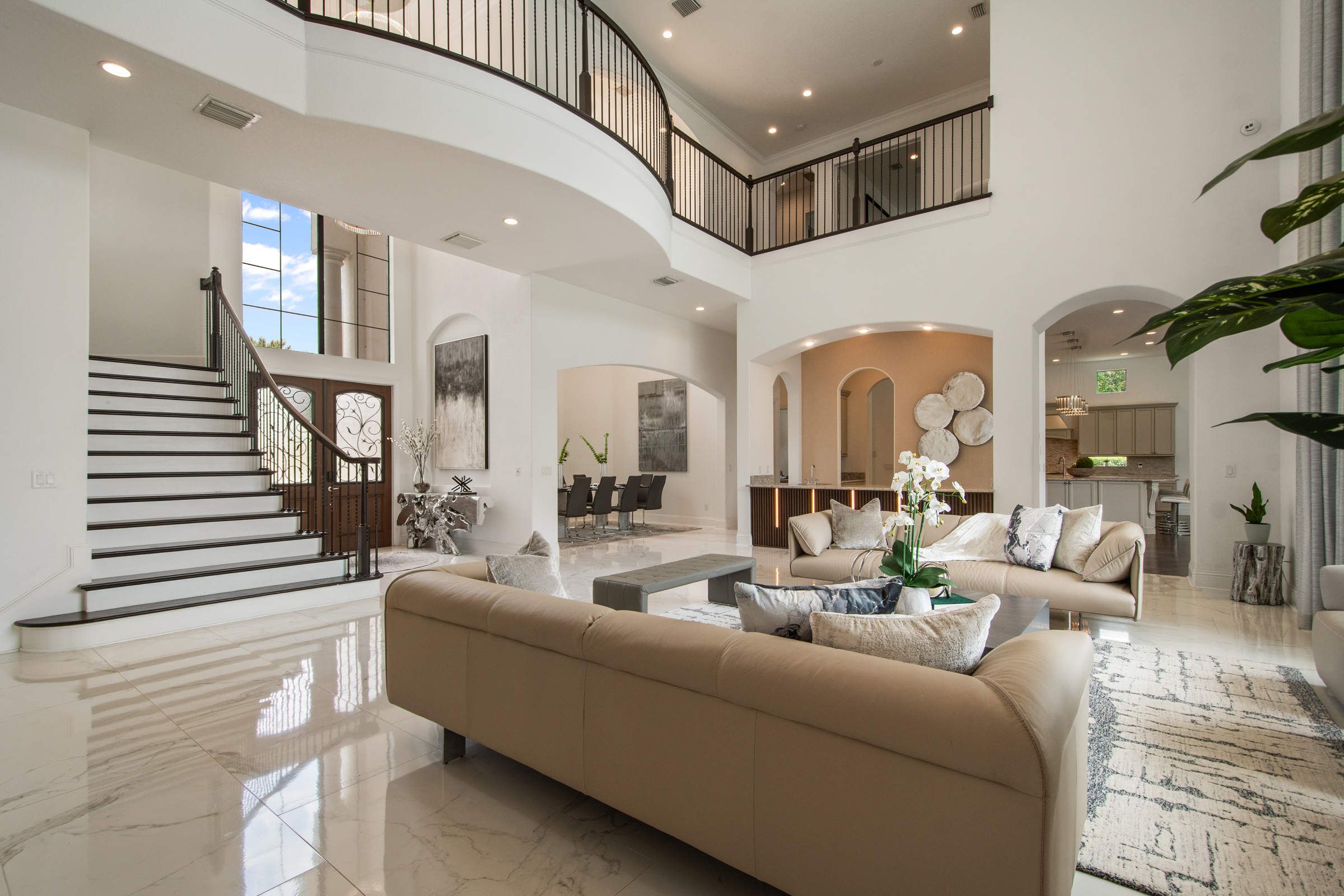 Spacious modern living room with beige sofas, marble floor, tall windows, and an open staircase—perfect for hosting an A-list gathering in September 2025. Dining room and kitchen are visible, with a neutral palette and natural light throughout.
