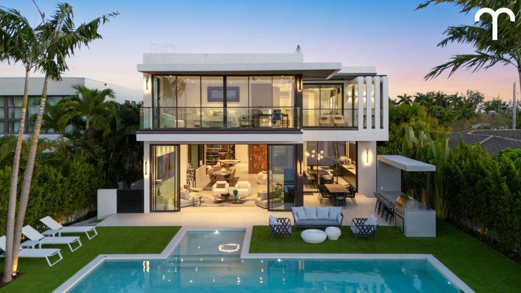 Modern two-story home with floor-to-ceiling glass windows, a balcony, outdoor pool, lounge chairs, barbecue area, and palm trees, set against a clear sky.