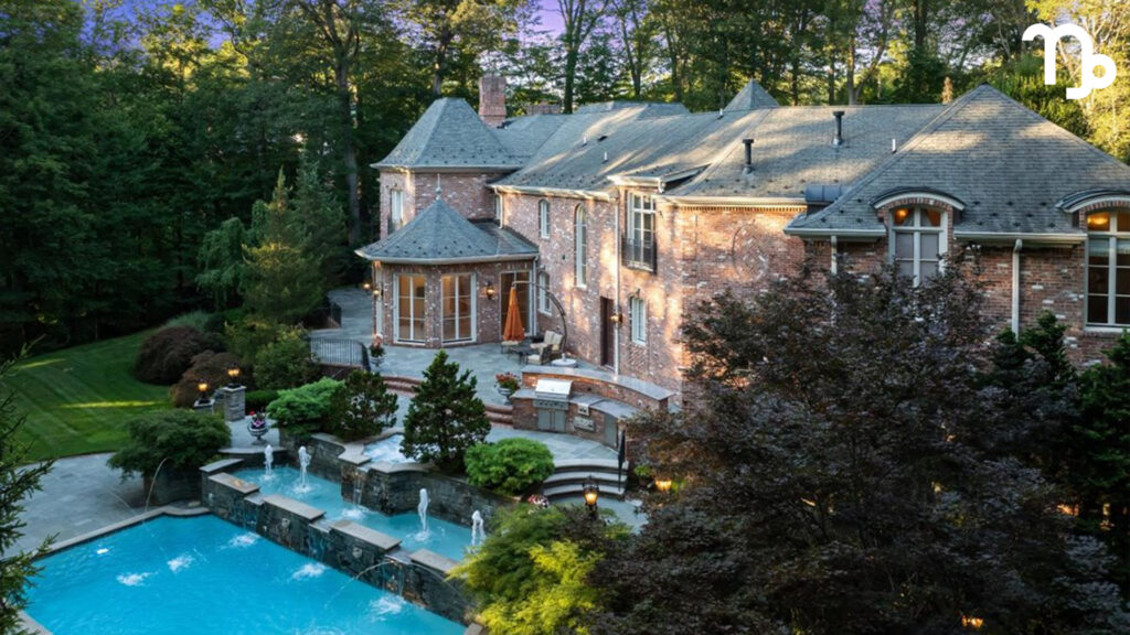 A large brick mansion with multiple windows and chimneys overlooks a landscaped backyard with a swimming pool, surrounded by trees and greenery. The Capricorn zodiac symbol in the top right hints at connections to astrological houses.