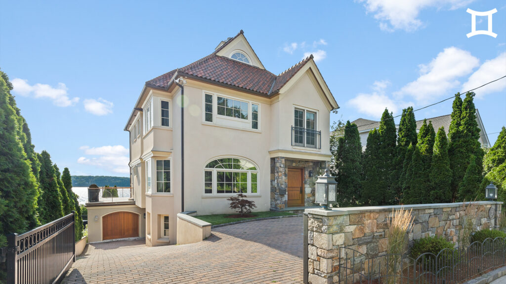 A beige two-story house with large windows, a tiled roof, and stone accents sits behind a gated driveway lined with tall evergreens, offering peaceful lake views—an ideal retreat for reflecting during Virgo season.
