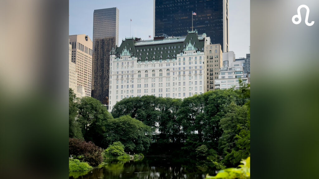 Cityscape featuring a historic white building with green roofs, surrounded by tall modern buildings, a lush park, and a reflective pond in the foreground.