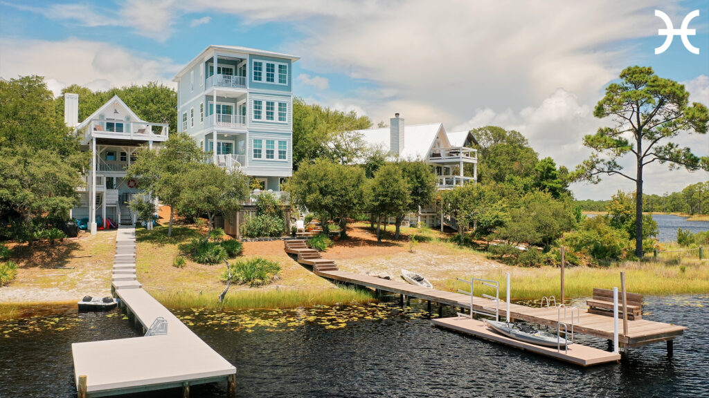Waterfront view of three light-colored multi-story homes with private docks, surrounded by trees, featuring a kayak and a partly cloudy sky over a calm body of water.