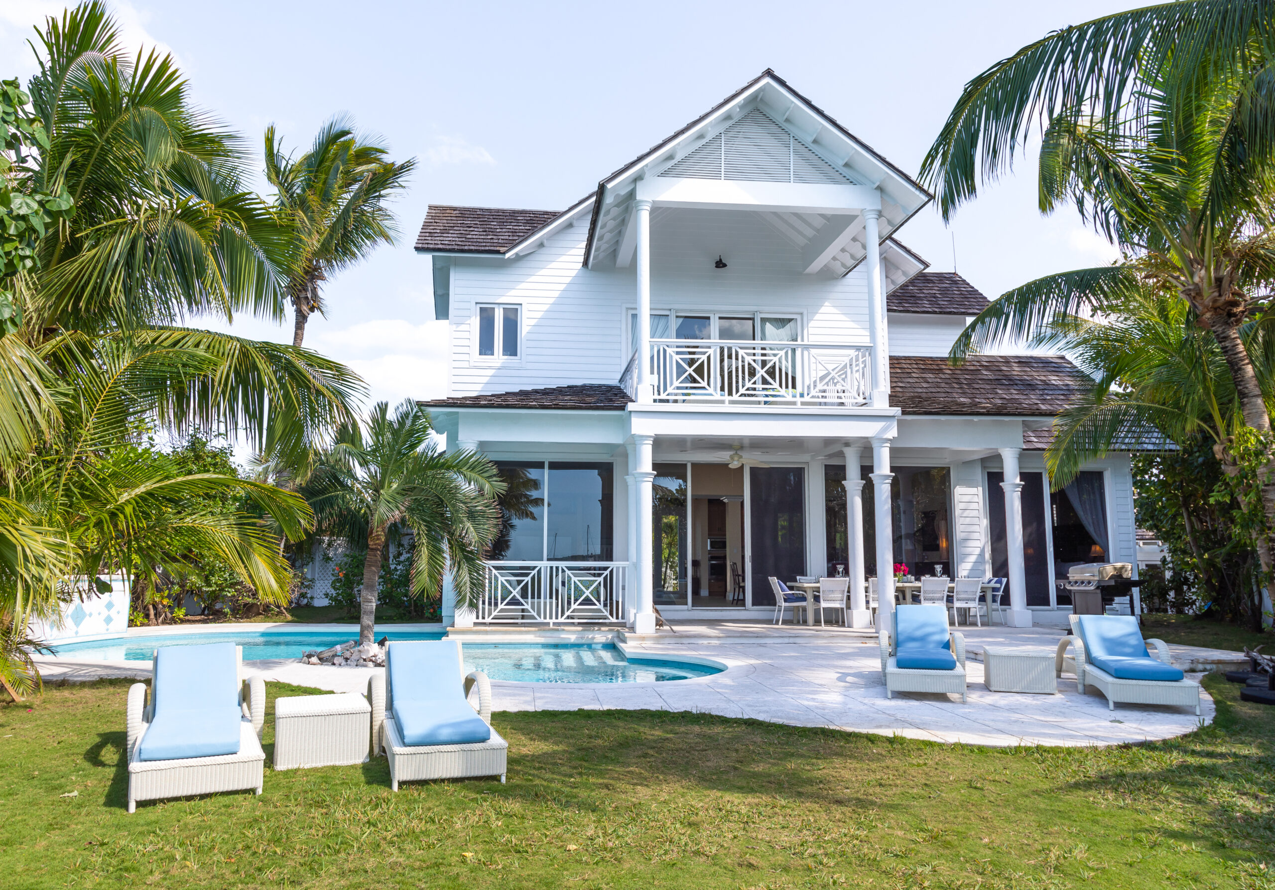 Exterior of Caribbean villa in the Bahamas, with a white porch, a kidney shaped pool exterior and pale blue lounge chairs sitting on the grass.