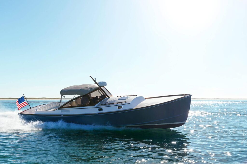 A motorboat with a canopy and an American flag cruises quickly across calm blue water, a stack of cookbooks visible on deck, as it creates a wake behind it under the clear sky.