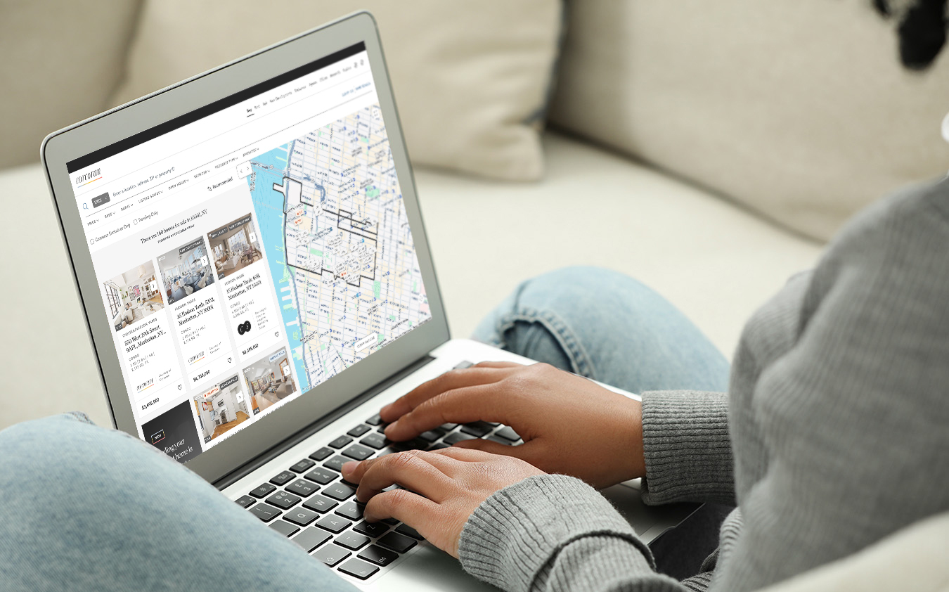 A person using a laptop sits on a couch, viewing a search result page of Manhattan home listings on Corcoran.com.