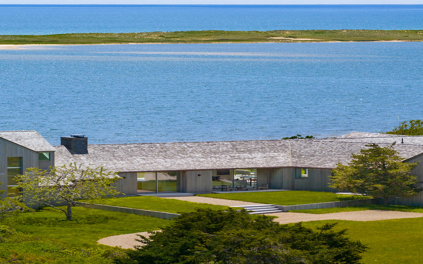On Martha's Vineyard, a modern house with a gray roof sits near a calm blue bay, with a sandy shoreline and grassy landscape in the background under a clear sky.