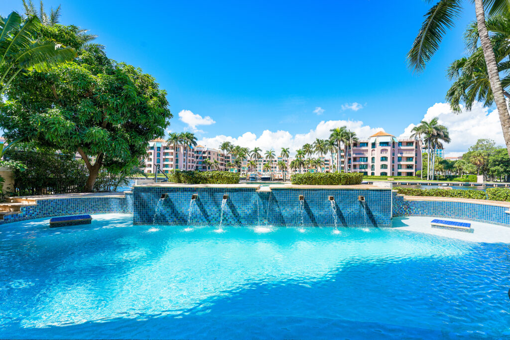 Swimming pool with cascading water spouts, surrounded by palm trees and greenery, with multi-story buildings in the background under a bright blue sky.