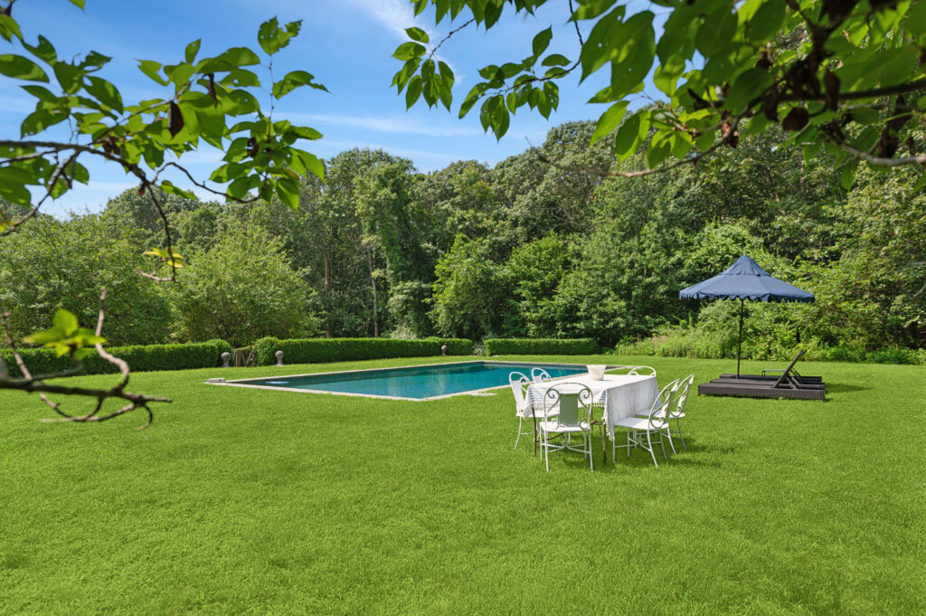 Rectangular backyard swimming pool surrounded by green grass, with a white outdoor dining set, shaded lounge chairs under a blue umbrella, and dense trees providing privacy.