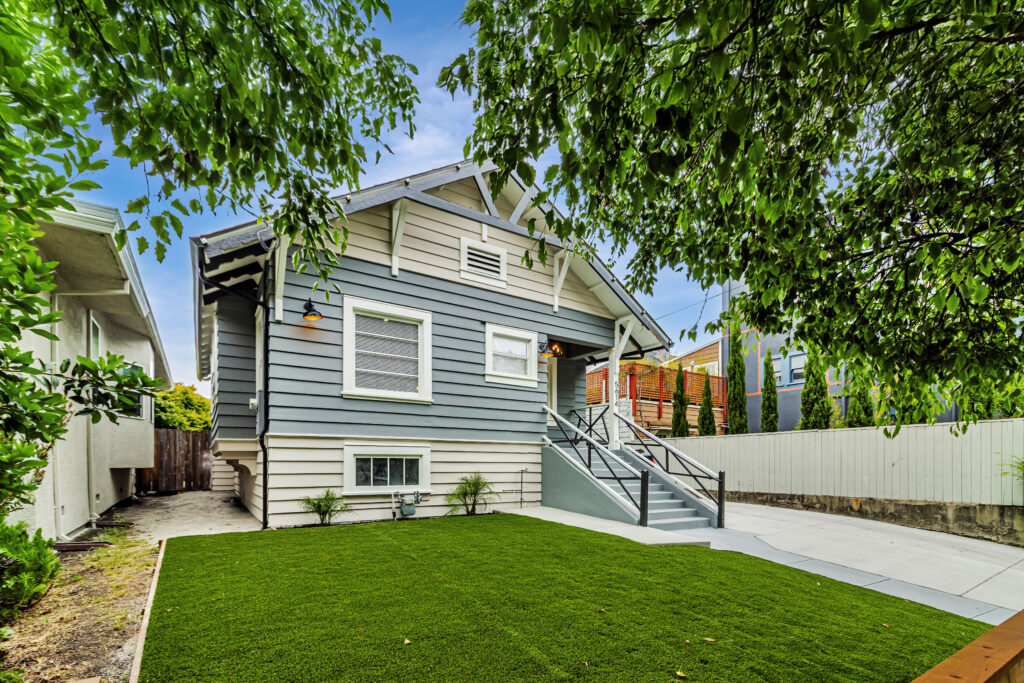 A gray craftsman-style house with white trim, a small porch with stairs, and a well-kept green lawn sits amid trees and neighboring homes in a charming college town.