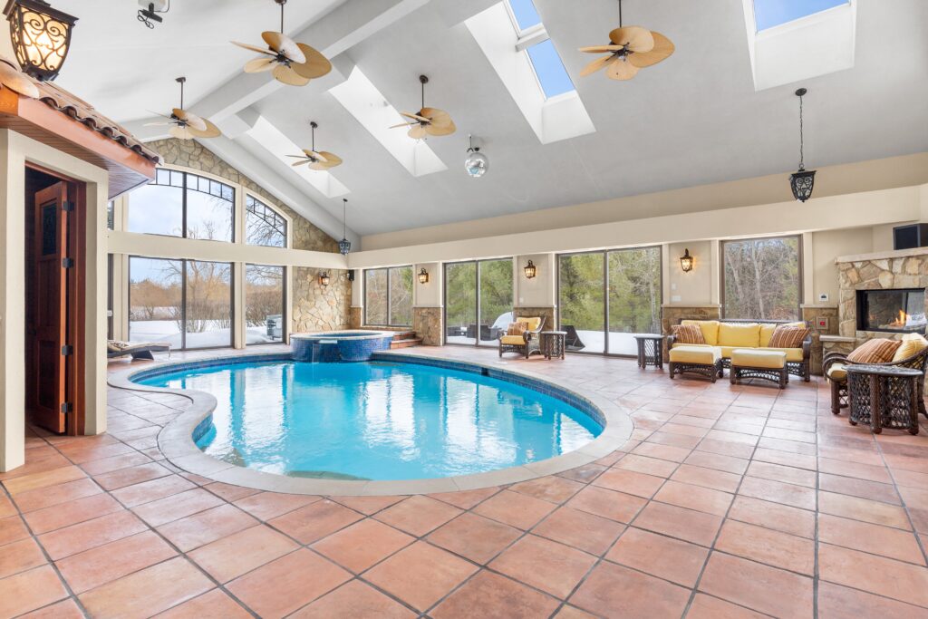 Indoor swimming pool with curved design, terracotta tile flooring, skylights, large windows, seating areas, and a cozy fireplace.