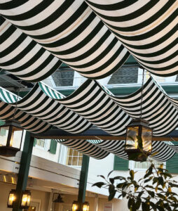 Green and white striped fabric awnings hang in waves from the ceiling of an indoor courtyard, with several lantern-style lights suspended beneath them and green window shutters visible in the background.