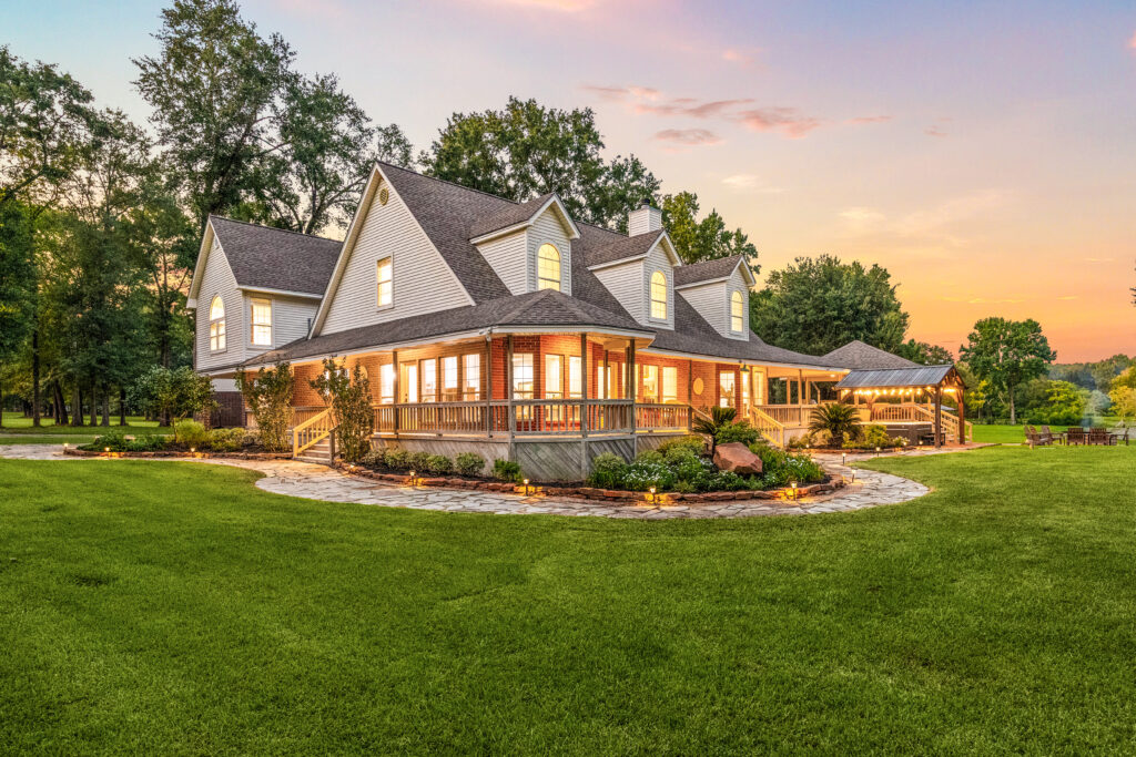 Large, modern farmhouse with a wraparound porch, lit windows, and manicured green lawn; surrounded by trees at sunset, with a gazebo or pavilion in the background.