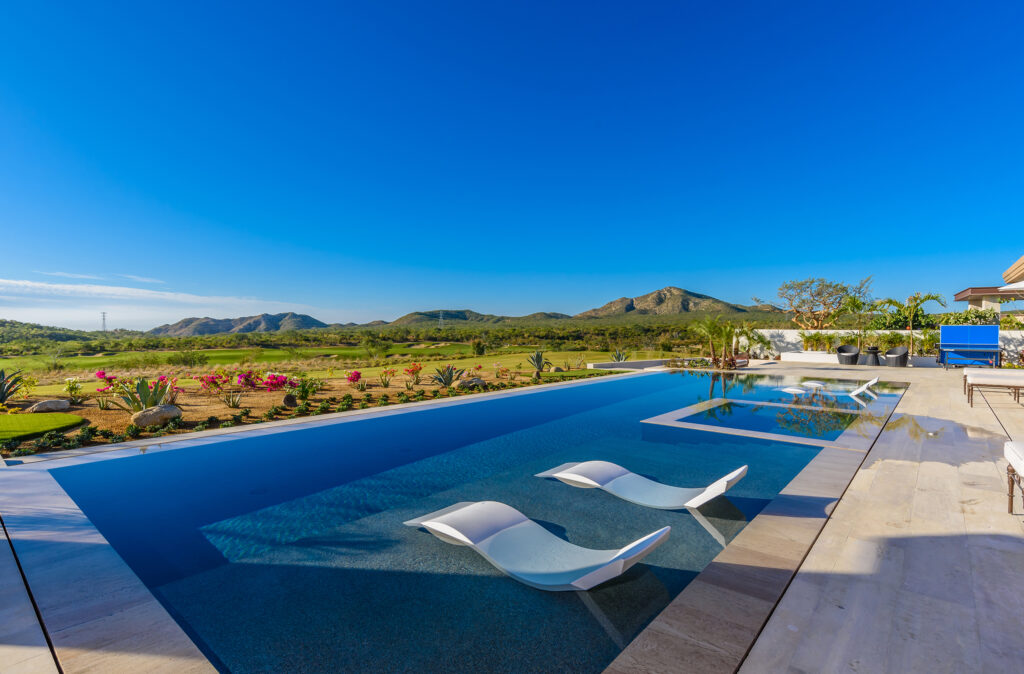 Luxurious outdoor swimming pool with in-water lounge chairs, surrounded by a vibrant garden and overlooking scenic green fields and mountains under a clear blue sky.