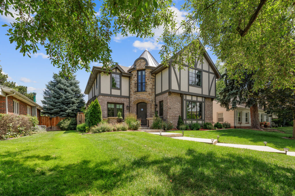 A two-story Tudor-style house with brick and timber accents sits under a blue sky with scattered clouds, surrounded by a green lawn, shrubs, and mature trees.