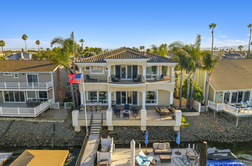 A waterfront two-story house with balconies, patios, palm trees, dock access, an American flag, and outdoor seating sits canal-side among similar homes under a clear blue sky.