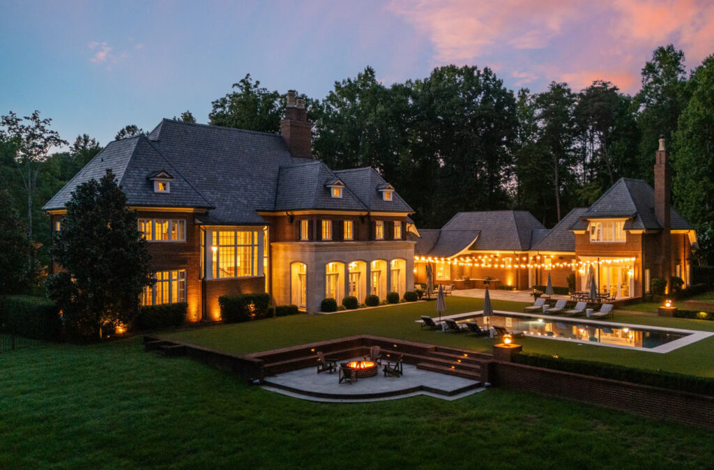 Elegant mansion at dusk with warm outdoor lighting, large windows, a swimming pool, and string lights over a patio.
