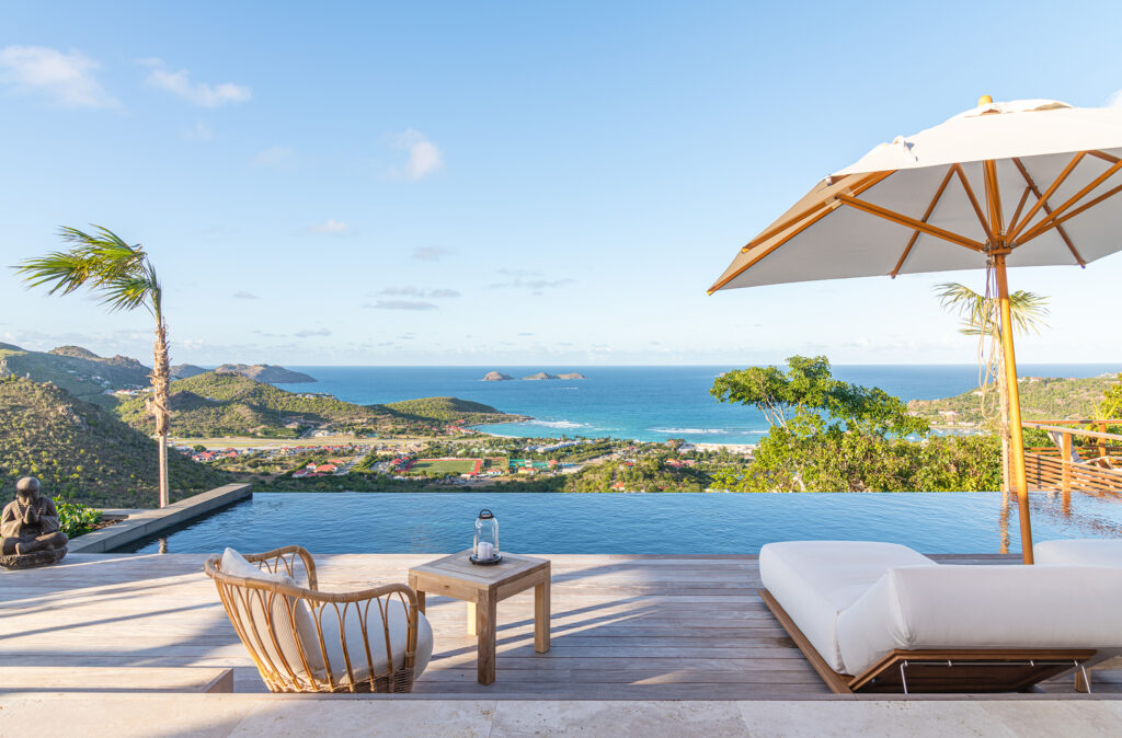Infinity swimming pool with ocean views, surrounded by a wooden deck, cushioned lounge chairs, a white umbrella, and coastal hills with scattered homes.