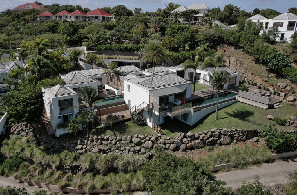 A modern white villa with multiple buildings, a pool, and lush greenery, situated on a hillside with stone landscaping.